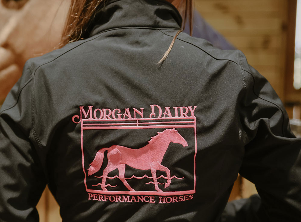 Una imagen de primer plano de una chaqueta negra con costuras rosas, con las palabras “Morgan Dairy Performance Horses”, usada por una mujer con cabello largo, liso y castaño.